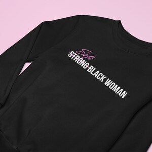 K&ouml;nnte beinhalten: Schwarzer Pullover mit wei&szlig;em Text, der "Soft Strong Black Woman" lautet, wobei das Wort "Soft" durchgestrichen ist. Das Wort "Soft" ist in Pink geschrieben.