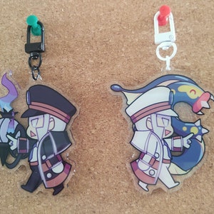 Könnte beinhalten: Zwei Acryl-Schlüsselanhänger mit stilisierten Anime-Figuren. Der linke Anhänger hat einen schwarzen Verschluss und zeigt eine Figur in dunkler Uniform mit einem geisterhaften Wesen. Der rechte Anhänger hat einen weißen Verschluss und zeigt eine Figur in weißer Uniform mit einem langen, blauen, aalartigen Wesen.
