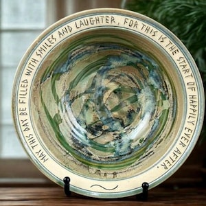 May include: A decorative ceramic bowl with a cream-colored rim and a green and blue interior. The bowl has the text "MAY THIS DAY BE FILLED WITH SMILES AND LAUGHTER, FOR THIS IS THE START OF HAPPILY EVER AFTER." The bowl is on a black stand.