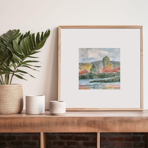 May include: A framed watercolor painting of a landscape with trees and a lake. The trees are in shades of green, yellow, and red, and the lake is a light blue. The sky is a light blue with a white cloud.