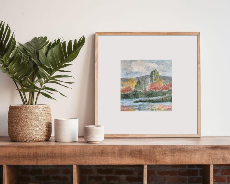 May include: A framed watercolor painting of a landscape with trees and a lake. The trees are in shades of green, yellow, and red, and the lake is a light blue. The sky is a light blue with a white cloud.