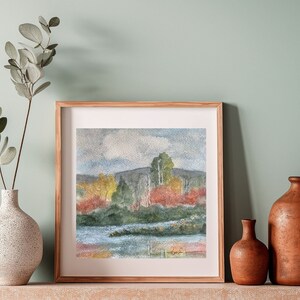 May include: A framed watercolor painting of a landscape with a lake, trees, and a cloudy sky. The painting is in a wooden frame and is hanging on a wall.