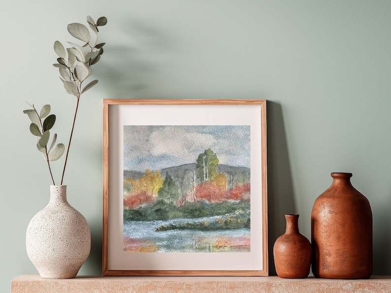 May include: A framed watercolor painting of a landscape with a lake, trees, and a cloudy sky. The painting is in a wooden frame and is hanging on a wall.
