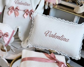May include: A white knitted baby outfit with a matching bonnet, a rectangular pillow with lace trim, and a knitted blanket. Each item features the name Valentina embroidered in pink thread. The outfit and blanket are accented with pink satin bows.