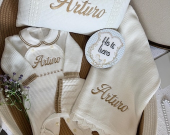 May include: A white quilted pillow, a knitted footed sleeper, a matching bonnet, and a knitted blanket, all embroidered with the name Arturo in gold thread. A circular sign with a blue checkered border reads He is here in black script.