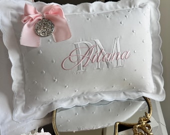 Puede incluir: Cojín rectangular blanco con borde festoneado y nombre "Aitana" bordado en rosa. Se adjuntan una cinta rosa y un dije plateado. El cojín tiene una textura punteada y se exhibe con un espejo decorativo y flores rosas.