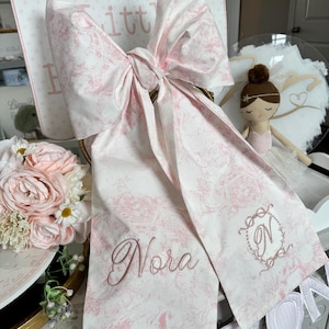 May include: A large, decorative bow made of white fabric with a pink floral pattern. The bow has long tails, with the name "Nora" embroidered in cursive. A monogram "N" is also embroidered. The bow is part of a baby or child's gift.