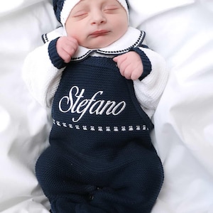Puede incluir: Un bebé recién nacido con un conjunto de punto azul marino y gorro a juego. El conjunto tiene el nombre "Stefano" bordado en blanco. El bebé está acostado sobre una sábana blanca.