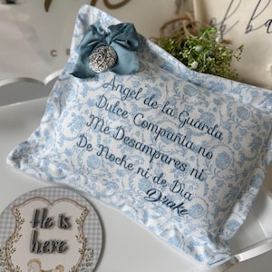 Op de afbeelding: Een decoratief kussen met een blauw bloemmotief en geborduurde Spaanse tekst. Het kussen is versierd met een blauwe strik en een zilveren ornament. De tekst luidt "Angel de la Guarda..." en "Drake".