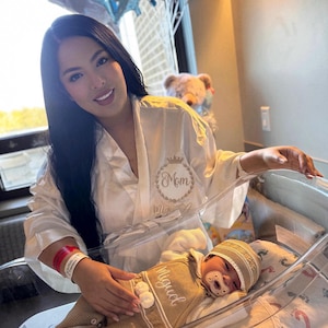 May include: A white satin robe with the word Mom and a crown graphic embroidered in gold, worn alongside a newborn baby in a clear bassinet. The baby wears a tan knitted romper with the name Miguel embroidered in white and a matching beanie.