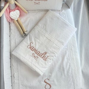 Pode incluir: Conjunto de roupa de cama para bebé branco com o nome "Samadhi" bordado. O conjunto inclui uma almofada, um cobertor e um lençol com elástico, todos com acabamento em renda. Uma boneca com tutu rosa também está no berço.