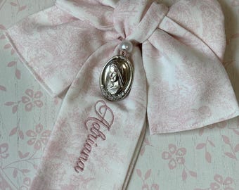Puede incluir: Un lazo de tela con estampado floral en rosa pálido y blanco que presenta un colgante ovalado de tono plateado con un retrato en relieve de una figura. El nombre Adriana está bordado en hilo rosa en una de las caídas del lazo, con una cuenta blanca.