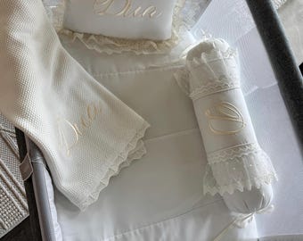 Puede incluir: Juego de ropa de cama para bebé blanco con bordado dorado "Dua". El juego incluye una almohada, una manta y un cojín cilíndrico, todos con encaje. La ropa de cama está colocada dentro de un cochecito gris.