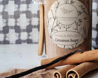 Könnte beinhalten: Ein Glasgefäß mit Bambusdeckel, gefüllt mit braunem, mit Vanille versetztem Zimtzucker. Das Gefäß trägt ein Etikett mit der Aufschrift "Epicurious Infusions, Locally Made, Locally Loved, Vanilla Infused Cinnamon Sugar". Auf einer weißen Oberfläche liegen drei Zimtstangen und eine Vanilleschote mit einer Prise Zimtzucker.