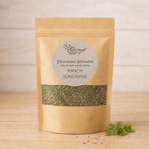 May include: A brown kraft paper pouch containing ranch seasoning, featuring a circular label with the text Epicurious Infusions, Locally Made, Locally Loved, and Ranch Seasoning, with a clear window displaying the green and white herb blend.