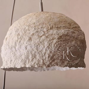 May include: A beige, textured pendant light with a rounded, dome-like shape. The light fixture has a rough, organic appearance and hangs from a thin, gray cord. The bottom edge has a slightly frayed texture. The logo "KRYSTYNA KRAWIEC ART DESIGN" is visible.