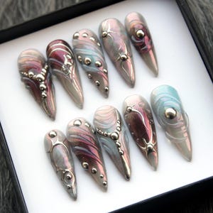 May include: Set of ten long, pointed, iridescent false nails with silver and white designs. The nails have a pearlescent finish and feature swirls, dots, and lines.