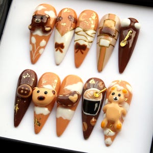 May include: A set of ten almond-shaped acrylic nails with various designs in shades of brown, beige, and white. Some nails feature 3D teddy bears, bows, and gold accents. The designs include coffee, ice cream, and other dessert-themed elements.