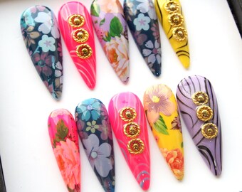 Op de afbeelding: Een verzameling van tien lange, amandelvormige kunstnagels. De nagels hebben verschillende ontwerpen, waaronder bloempatronen, effen kleuren en gouden versieringen. Kleuren zijn onder meer roze, blauw, geel en paars.