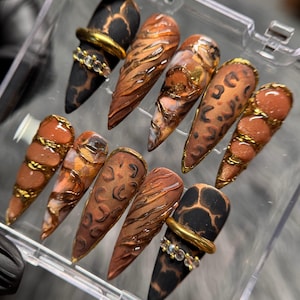 May include: A set of ten stiletto-shaped press-on nails featuring a mix of matte black leopard print, metallic copper textures, and glossy brown finishes. Some nails are adorned with gold-toned rings and small clear rhinestones.