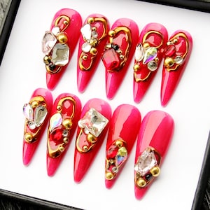 May include: A set of ten long, pointed artificial nails in a vibrant pink hue. Each nail is adorned with gold-colored embellishments, sparkling rhinestones, and small gold beads, creating a glamorous and ornate design. The nails are arranged neatly in a row.