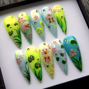 May include: Ten long, pointed, fake nails with a light blue, green, and yellow base. Each nail is decorated with a different design featuring a small yellow chick, green leaves, and pink flowers.