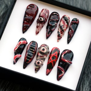 May include: Set of ten false nails with various spooky designs, including a snake, an eye, a pentagram, and a bloody stripe. The nails are painted in shades of black, red, and white.
