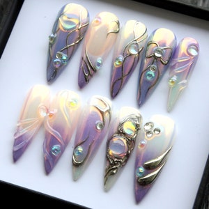 May include: Set of ten iridescent purple and white false nails with intricate gold and silver designs, including bows, swirls, and gems.