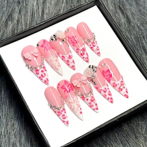 May include: A set of ten pink and white stiletto-shaped false nails. Each nail features a pink base with white tips, floral designs, and leopard print accents. Some nails have bows and rhinestone embellishments.