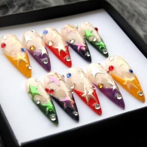 May include: Set of ten false nails with a white base and a gradient of color on the tips. The tips are painted in various colors, including red, green, purple, and blue. Each nail is decorated with gold stars and rhinestones.