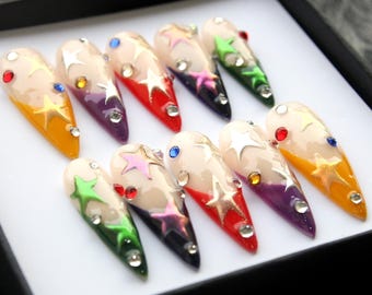 Puede incluir: Conjunto de diez uñas postizas con una base blanca y un degradado de color en las puntas. Las puntas están pintadas en varios colores, incluyendo rojo, verde, morado y azul. Cada uña está decorada con estrellas doradas y pedrería.