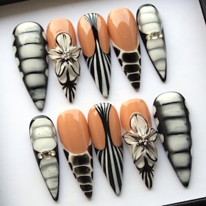 May include: A set of ten long, almond-shaped artificial nails. Designs include black and white patterns, peach-colored bases, and 3D flower accents. Some nails feature rhinestone embellishments.