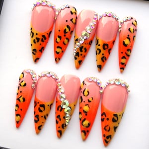 Puede incluir: Un conjunto de diez uñas artificiales largas en forma de almendra. Cada uña presenta un vibrante diseño de estampado de leopardo naranja y amarillo, con una base rosa y una línea de brillantes pedrería a lo largo del borde o en el centro.