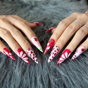 May include: Stiletto-shaped press-on nails featuring a mix of designs including red and pink floral patterns, leopard print accents, and small ladybug illustrations. The set displays a vibrant color palette of bright red, soft pink, and black.