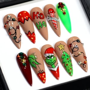 May include: Set of ten Christmas-themed false nails with various designs, including a Grinch, a Christmas tree, a candy cane, and lights. The nails are nude with red, green, and white accents.