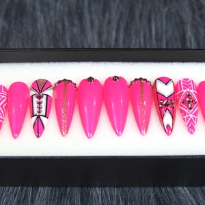 Puede incluir: Conjunto de diez u&ntilde;as postizas stiletto rosas con dise&ntilde;os geom&eacute;tricos blancos y dorados y pedrer&iacute;a.