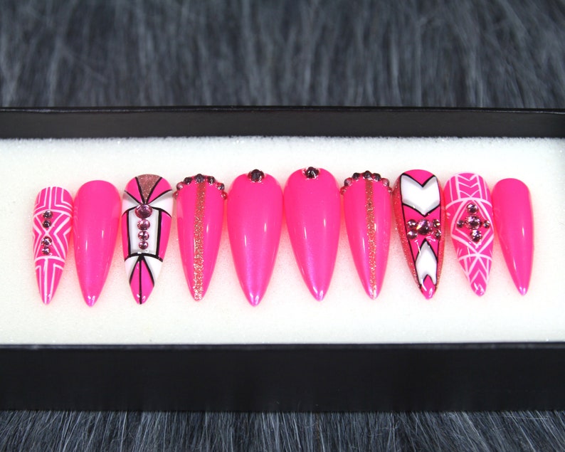 Puede incluir: Conjunto de diez u&ntilde;as postizas stiletto rosas con dise&ntilde;os geom&eacute;tricos blancos y dorados y pedrer&iacute;a.