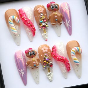 May include: A set of ten long, almond-shaped artificial nails with a Christmas theme. Designs include gingerbread men, candy canes, Christmas trees, snowflakes, and pink and white accents. The nails are decorated with glitter and rhinestones.