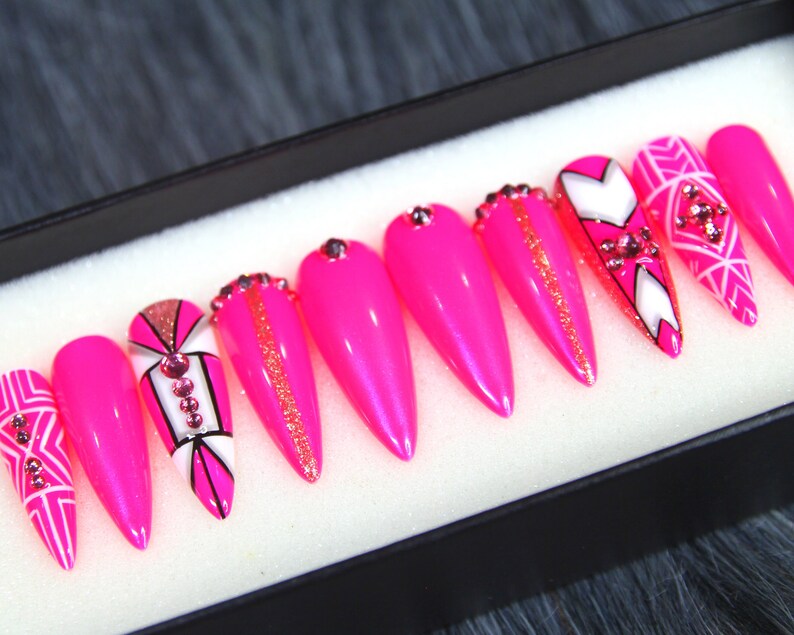 Puede incluir: Conjunto de diez u&ntilde;as postizas stiletto rosas con dise&ntilde;os geom&eacute;tricos blancos y negros y pedrer&iacute;a.