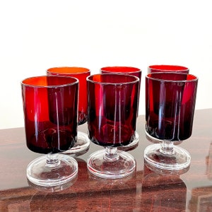 Könnte beinhalten: Sechs Vintage-Weingläser mit klarem Stiel und Boden sowie tiefroter Schale. Die Gläser sind auf einer dunklen Holzoberfläche angeordnet. Das Innere der Gläser hat einen orangen Farbton.