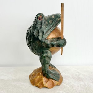May include: A handcrafted frog figurine with a dark green body and red eyes, holding a wooden stick. The frog is perched on a brown, textured base. The figurine has a rustic, aged appearance, suggesting it is vintage or antique.