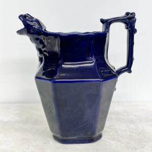 May include: A deep cobalt blue ceramic pitcher featuring a hexagonal body and a glossy finish. The spout is molded into the shape of a stylized animal head with an open mouth, and the handle is designed to resemble a textured, knotted branch.