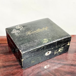 May include: A black leather box with a gold inscription that reads "Handkerchief" and a silver monogram "H.D." on the top. The box has a keyhole and is slightly worn.