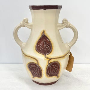 Könnte beinhalten: Eine weiße Keramikvase mit braunen Blattmotiven und zwei Henkeln. Die Vase hat eine strukturierte Oberfläche und einen braunen Rand.