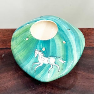 May include: A green and white ceramic vase with a horse design. The vase has a square base and a round opening at the top. There are several holes around the opening.