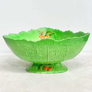 Könnte beinhalten: Eine hellgrüne Keramik-Servierschüssel in Form eines Kohlblattes. Die Schüssel hat einen Sockel und ist mit orangefarbenen und grünen Akzenten verziert. Die Schüssel ist mit einem strukturierten Blattmuster versehen.