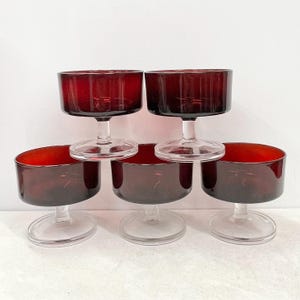 May include: Five vintage cocktail glasses in a deep burgundy hue. Each glass features a short, cylindrical bowl atop a clear, stemmed base. The glasses are arranged in a stacked formation, highlighting their design and colour.