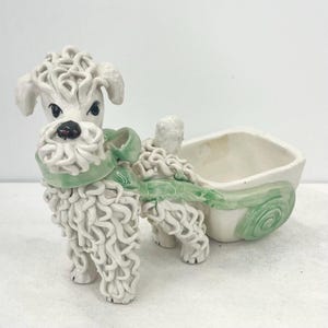 May include: A white ceramic poodle figurine with green details pulling a green and white ceramic planter. The poodle has curly hair and a green ribbon around its neck.