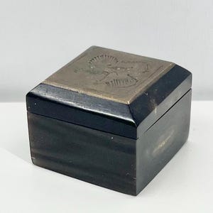 Könnte beinhalten: Eine kleine, quadratische, schwarze Box mit einem silbernen Deckel. Der Deckel hat ein kompliziertes Design eines Vogels oder Tieres.