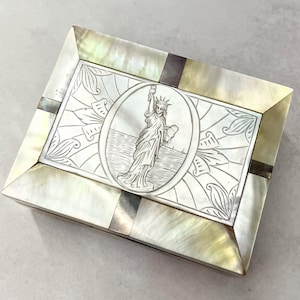 以下が含まれることがあります： 中央に自由の女神の浮き彫りがある長方形の真珠貝の箱。箱は花柄で飾られ、濃い茶色の縁取りが施されています。